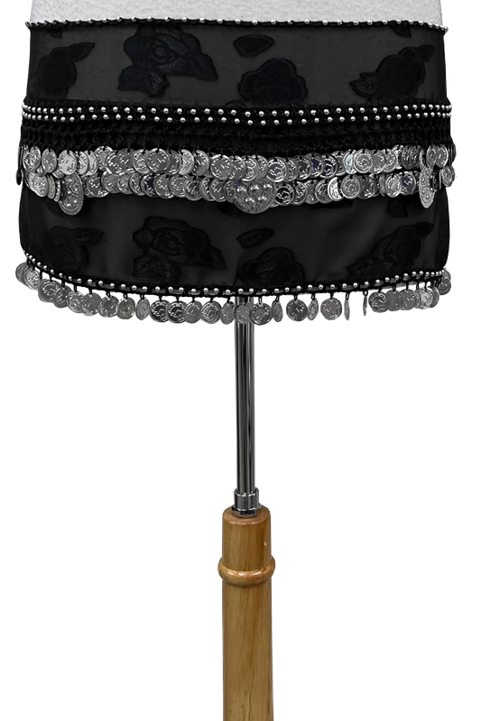 A black fabric hip sash with floral patterns, silver coin fringe, and bead trim, displayed on a dress form stand against a white background.