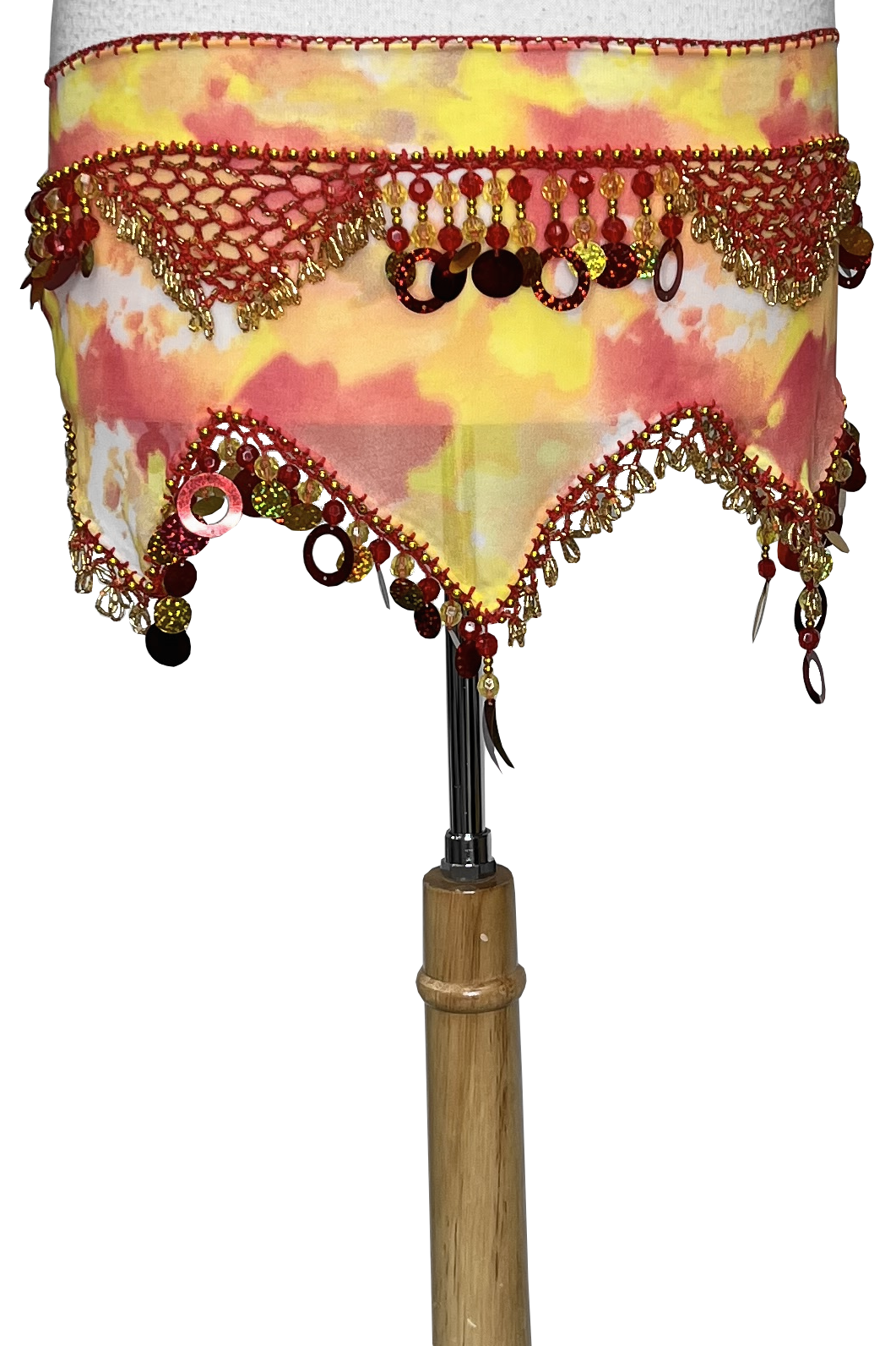 A close-up of a belly dance skirt draped over a wooden dress form, featuring a yellow and pink watercolor pattern with red and gold beaded fringe and tassels.