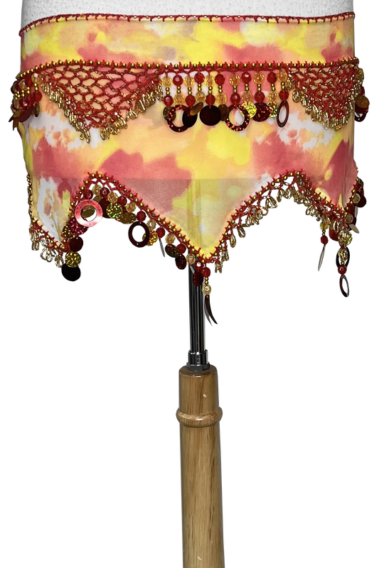 A close-up of a belly dance skirt draped over a wooden dress form, featuring a yellow and pink watercolor pattern with red and gold beaded fringe and tassels.