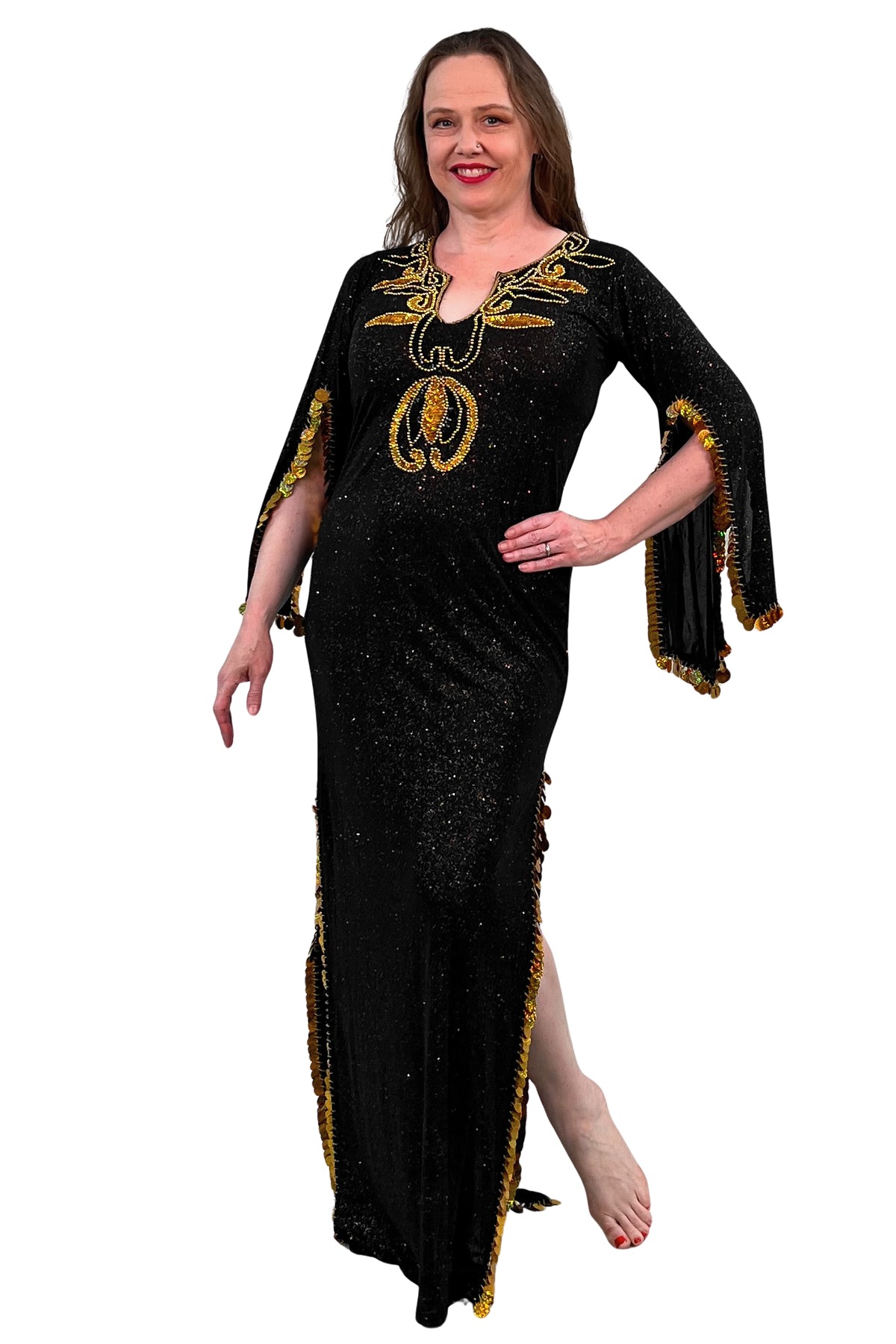 A woman modeling a black floor-length embellished dress with gold sequins, draped sleeves, and gold fringe trim against a plain white background.