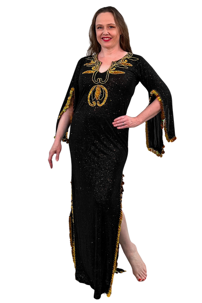 A woman modeling a black floor-length embellished dress with gold sequins, draped sleeves, and gold fringe trim against a plain white background.