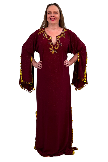 A woman modeling a long burgundy maxi dress with gold sequins and paillette trim on the sleeves and hem, set against a plain white background.