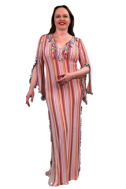 A woman modeling a long, sleeveless maxi dress with vertical orange, pink, white, and red stripes, featuring colorful embroidered details on the neckline and fringed trim on the sleeves and sides.