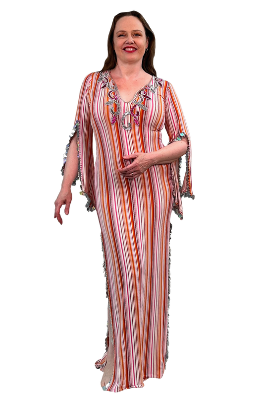 A woman modeling a long, sleeveless maxi dress with vertical orange, pink, white, and red stripes, featuring colorful embroidered details on the neckline and fringed trim on the sleeves and sides.