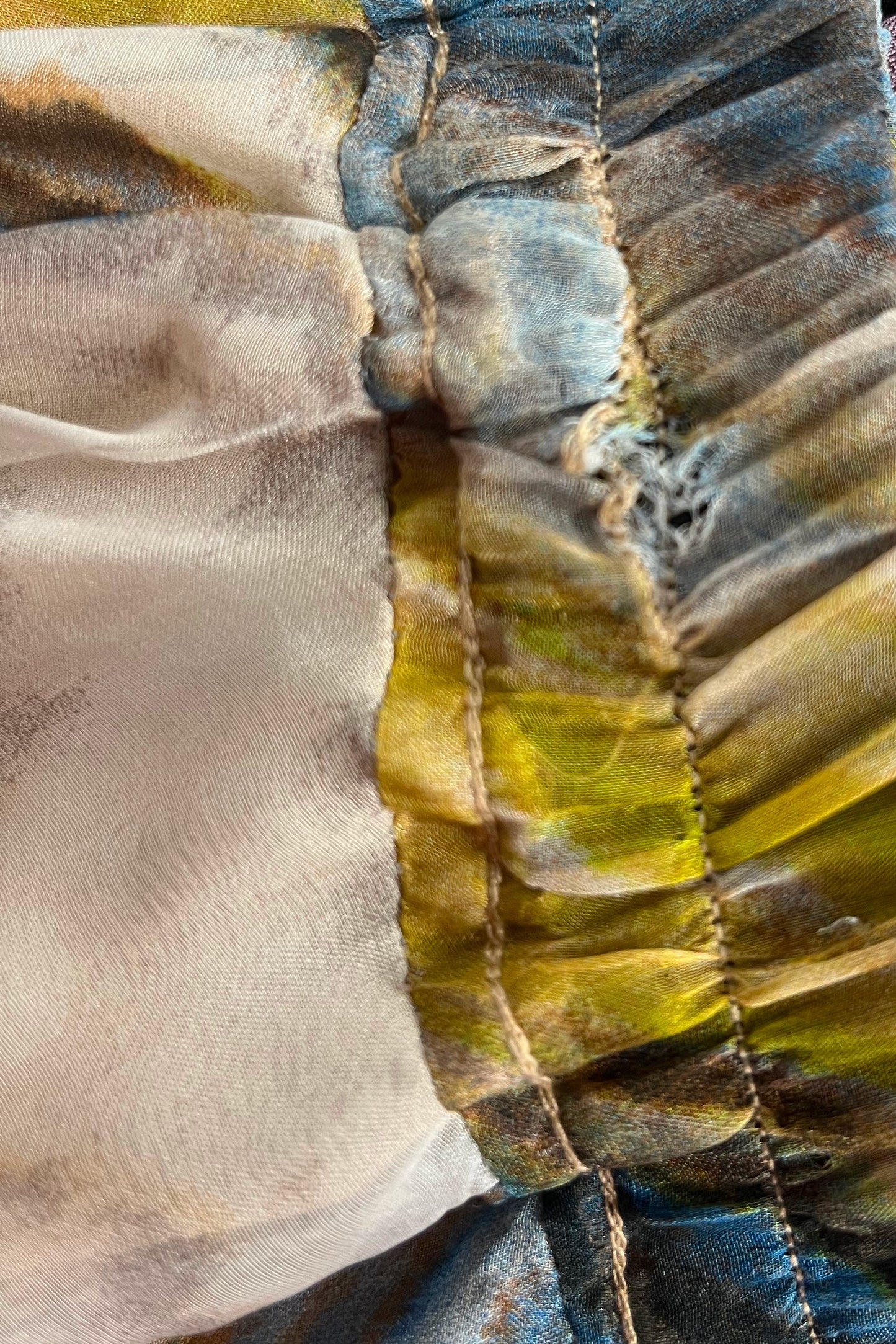 Close-up of a ruffled garment detail showing a sheer beige fabric panel joined to a pleated section with blue, olive green, and brown tie-dye or batik-style patterning.