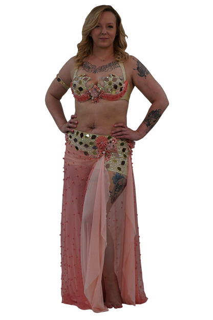A woman modeling a belly dance costume consisting of a gold and pink embellished bra top, a matching sheer pink skirt, and  gold sequined shorts, set against a plain white background.